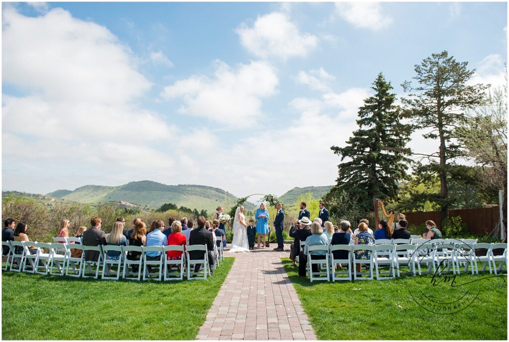 a view from the back of the wedding ceremony shows people seated in white chairs as the couple gets married with a backdrop of green foothills and blue partly-cloudy skies.