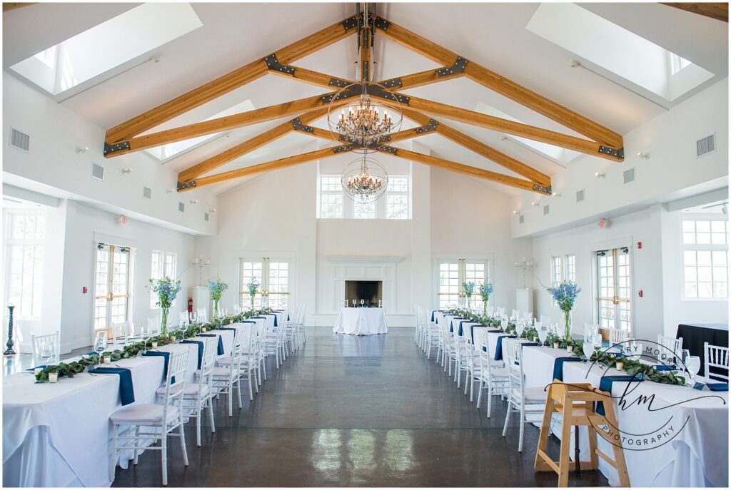 Two room-length dinner tables run on both sides of a long white reception hall decorated with industrial-themed wooden beams.