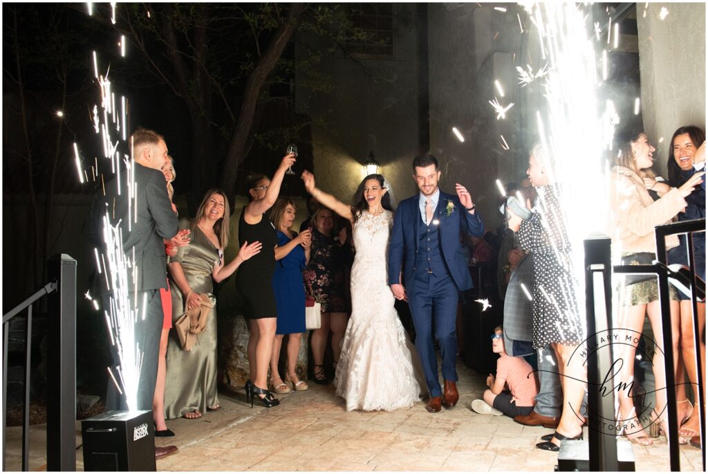 The bride and groom walking out while firework fountains spray white sparkles after getting married.